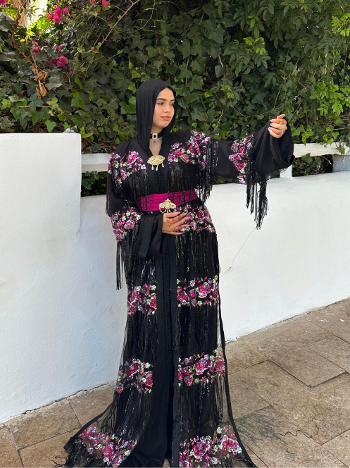 caftan élégant noir orné de broderies florales violettes et roses, avec des franges décoratives et une ceinture fuchsia.